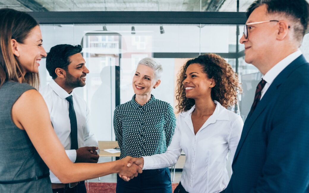 Two women shaking hands with a few other people surrounding them
